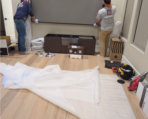 Technicians installing a large projection screen in a dedicated home theater space with equipment staged below.
